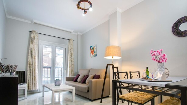 A modern living room with a sofa, a coffee table, a dining table with chairs, a lamp, and a vase with flowers.