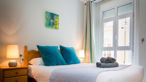 A neatly made bed with a blue blanket, two teal pillows, and a rolled towel on the side. There is a wooden nightstand with a lamp and a small drawer. A colorful abstract painting hangs on the wall above the bed.