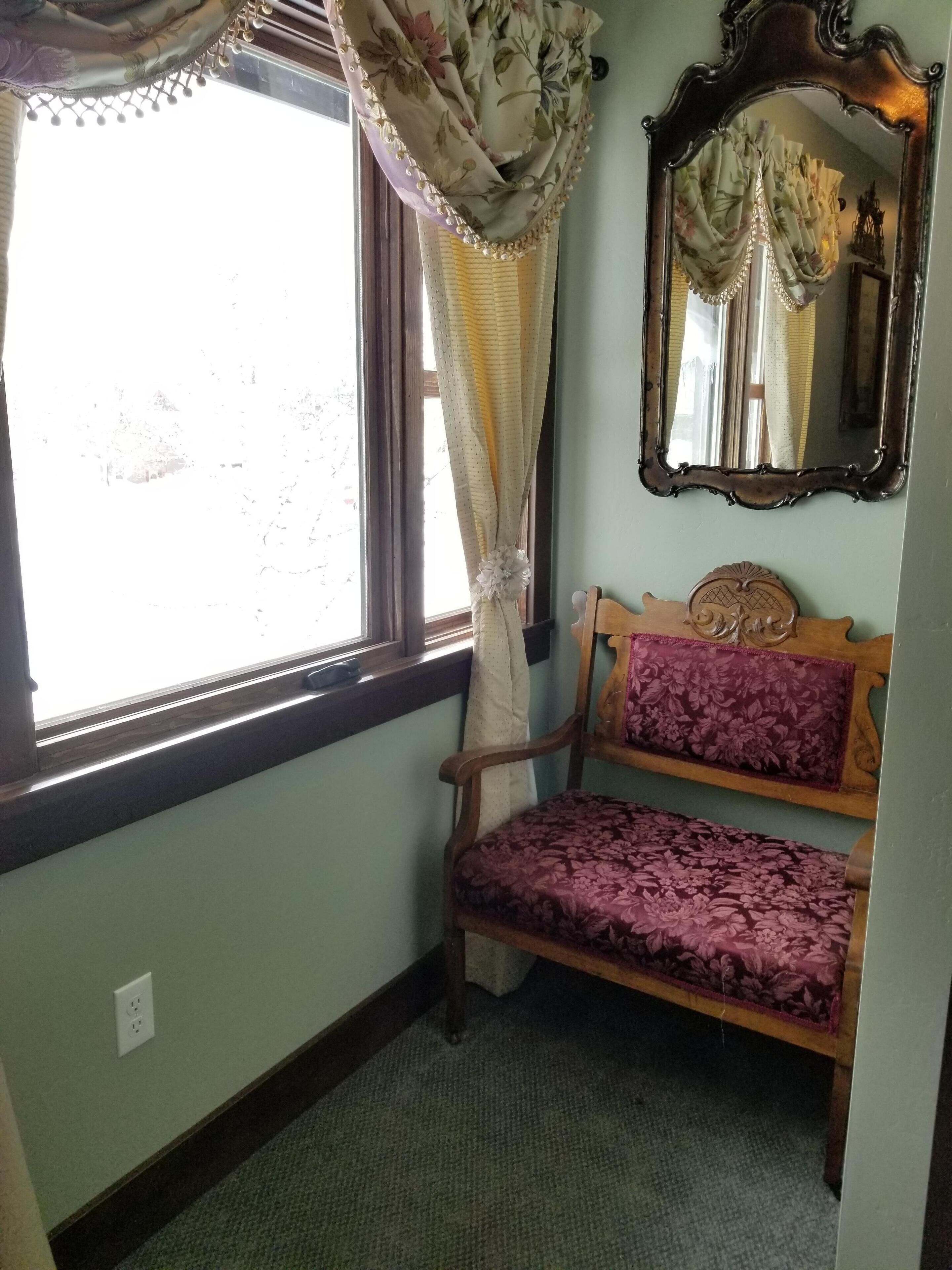 A room with a window, a wooden bench with floral upholstery, and a mirror with a floral curtain.
