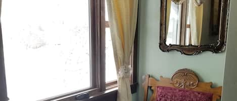 A room with a window, a wooden bench with floral upholstery, and a mirror with a floral curtain.