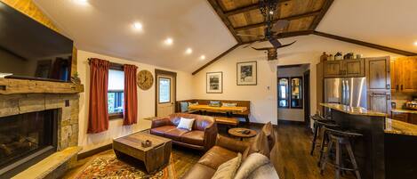 A spacious living room with a stone fireplace, leather sofas, a wooden ceiling, and a kitchen area with stainless steel appliances.