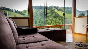 A modern living room with a large window offering a view of green hills. There is a brown sofa and a small ottoman. - Pousada Espelho DÁgua (Gonçalves)
