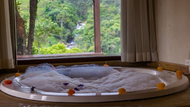 A hot tub with bubbles, surrounded by candles and flowers, with a view of a lush forest through a window. - Pousada Espelho DÁgua (Gonçalves)