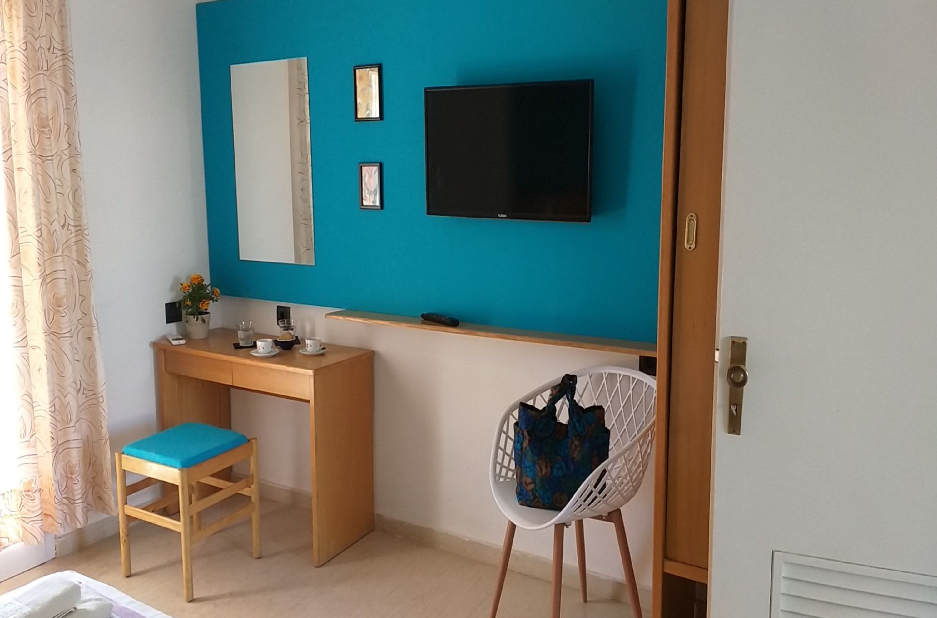 A room with a teal accent wall, a wooden desk with a chair, a television mounted above the desk, and a white chair with a patterned bag.