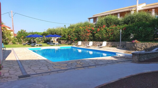 Una piscina con sillas de descanso y sombrillas, rodeada de vegetación y un muro de piedra.
