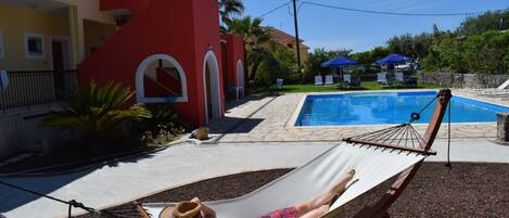 A hammock by a pool with a red building and a clear sky.