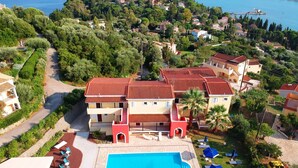 Un edificio grande con piscina y varios balcones, rodeado de abundante vegetación y vistas al mar.