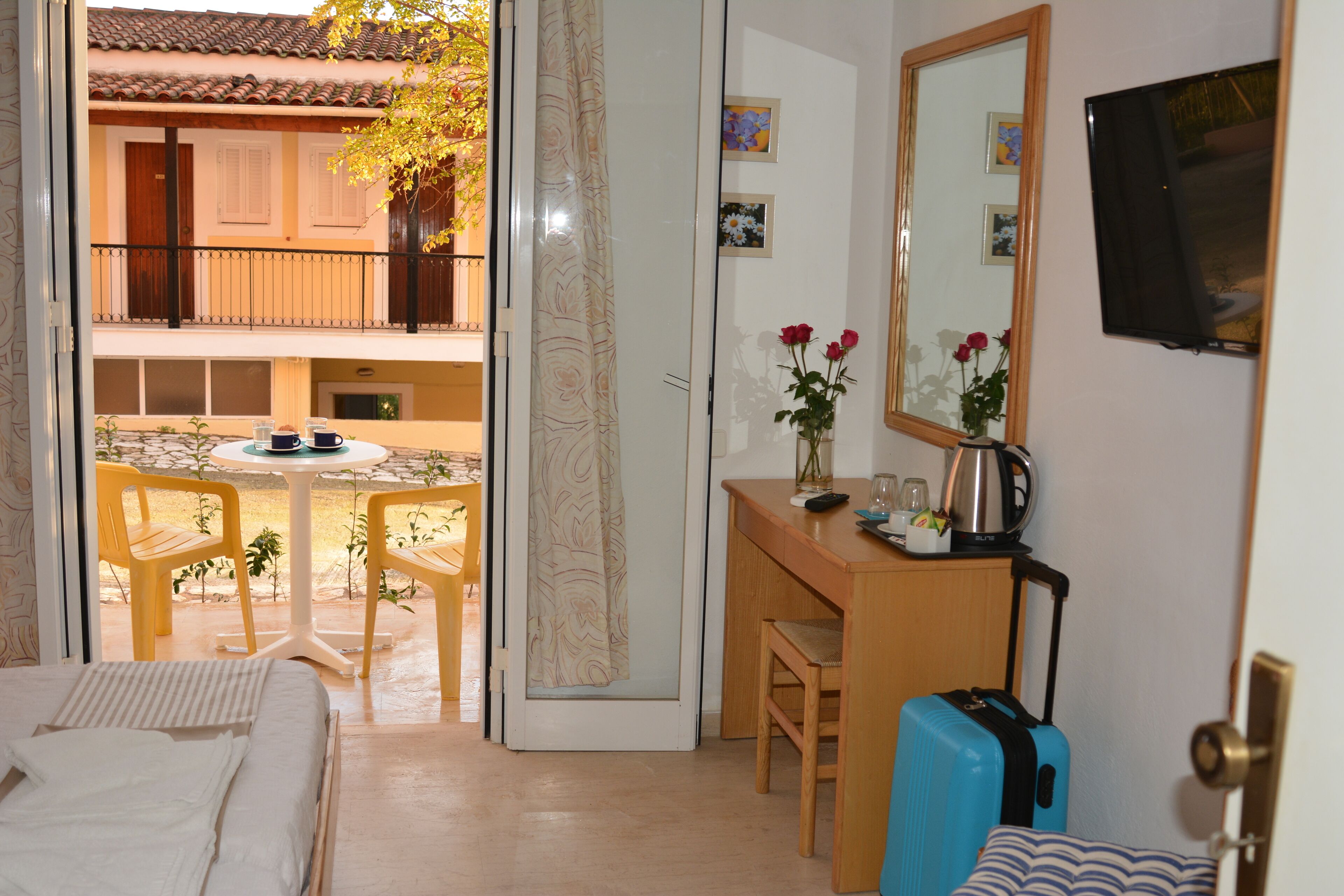 A hotel room with a bed, a desk with a chair, a small table with a chair, a window, and a balcony with a table and chairs.