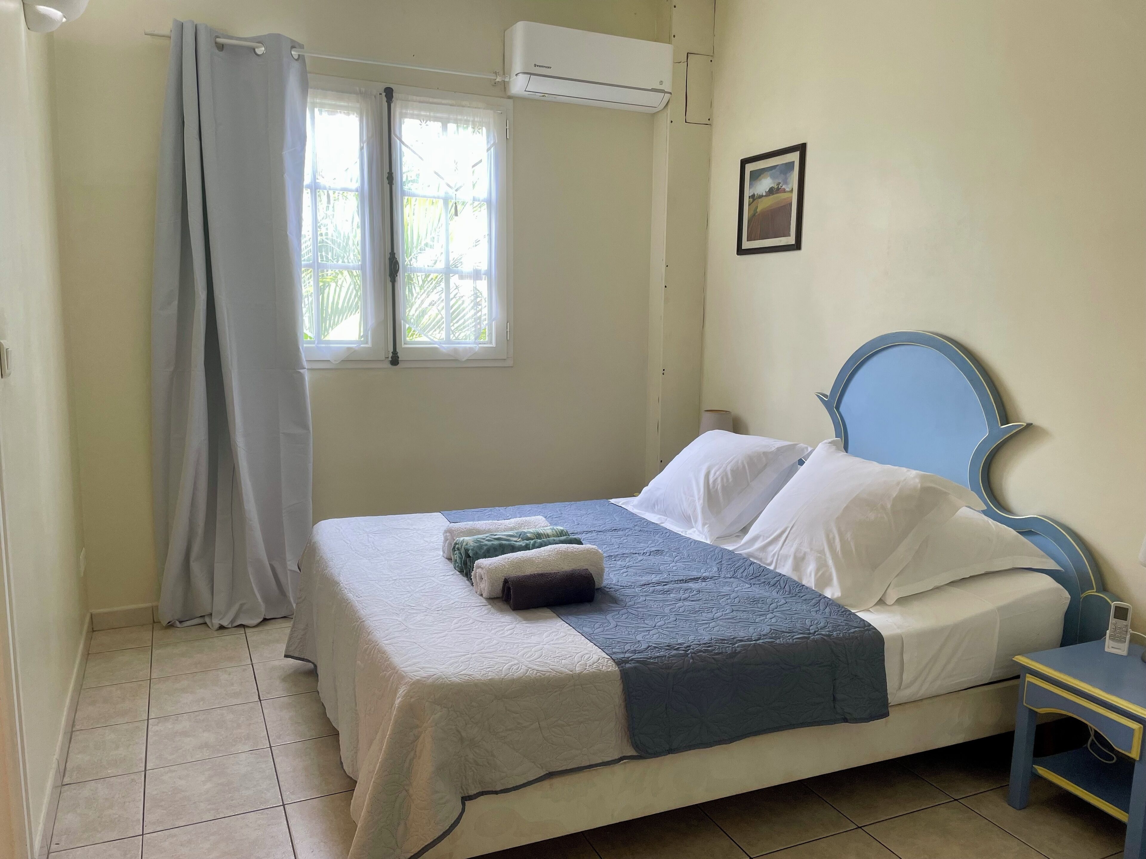 Un dormitorio con cama, mesita de noche, ventana con cortinas, aire acondicionado y un cuadro en la pared.