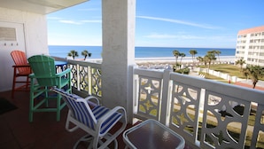 Ein Balkon mit Stühlen und einem Tisch, der auf einen Strand mit Palmen und ein Gebäude in der Ferne blickt.