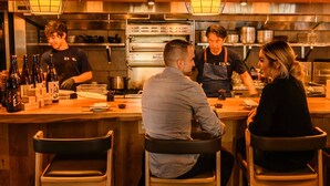 Una cocina de restaurante con cocineros preparando alimentos, una zona de bar con botellas y un área de comedor con sillas.