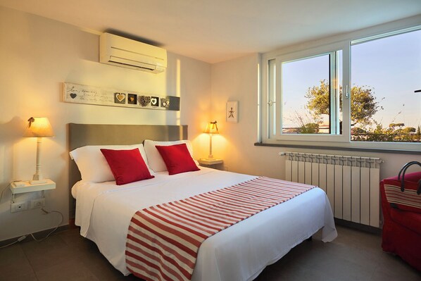 A neatly made bed with red pillows, a bedside lamp, and a window with a view of greenery. - Amazing penthouse with terrace, parking u.r. (Roma)
