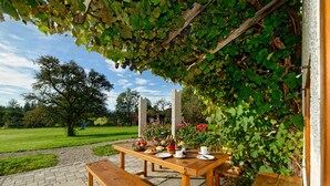 An outdoor wooden table set for a meal, surrounded by greenery and a clear sky.
