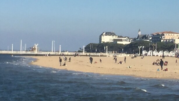 Een strand met een groot gebouw op de achtergrond, een pier met witte palen, en mensen op het zand.