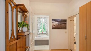 Ein Flur mit einem Holzschrank, einem Spiegel, einem Topfpflanzen, einem Fenster mit Vorhängen und einem Bett mit Blick auf Bäume.