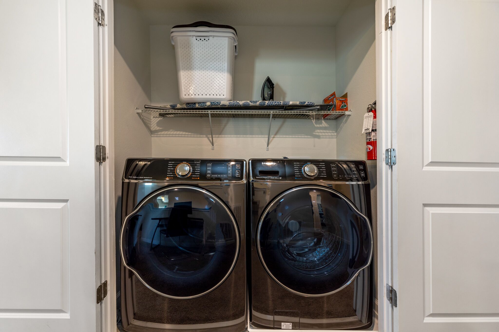 A laundry room with two front-loading washing machines, a drying rack, and a shelf with various items.