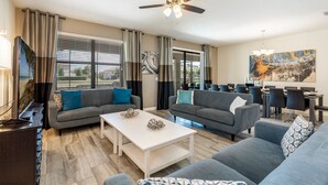 Ein modernes Wohnzimmer mit Deckenventilator, grauen Sofas, einem weißen Couchtisch und einem Essbereich mit einem großen Gemälde.