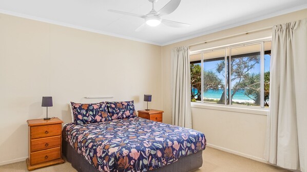 A bedroom with a bed, two bedside tables, a ceiling fan, a window with curtains, and a view of the beach.