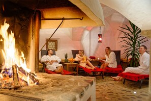Um grupo de pessoas relaxando perto de uma lareira em um quarto com piso de pedra e uma planta grande.