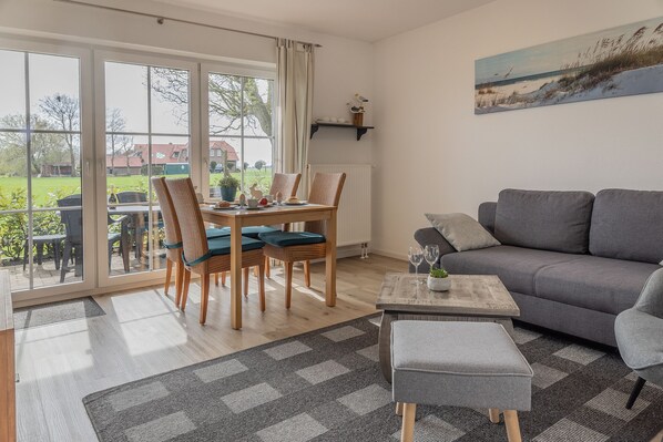 Un salon avec un canapé gris, une table à manger avec des chaises et une vue sur un jardin grâce à de grandes fenêtres.