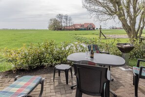 Une terrasse extérieure avec une table et des chaises, un petit jardin et une vue sur un grand champ verdoyant et une maison au loin.