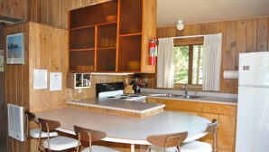 Una cocina con armarios de madera, estufa, pileta y refrigerador. Hay una mesa de comedor con sillas y un ventanal con cortinas.