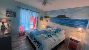 Et soveværelse med et maritimt muralmaleri, en seng med et surfbræt-design og et natbord med en lampe.