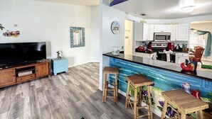 En stue med fjernsyn, en tv-reol i træ og et blåt natbord. Et køkken med hvide skabe og en sort bordplade ses i baggrunden.