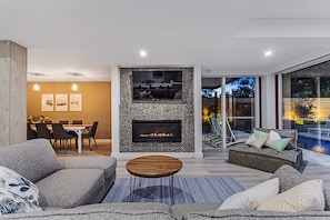 A modern living room with a fireplace, a dining area, and a view of a swimming pool. - La Luna Shoal Bay Luxury Holiday Home with Pool (Shoal Bay)