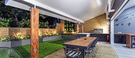 Un patio esterno moderno con un pavimento in legno, un tavolo da pranzo con sedie e una griglia per barbecue integrata.