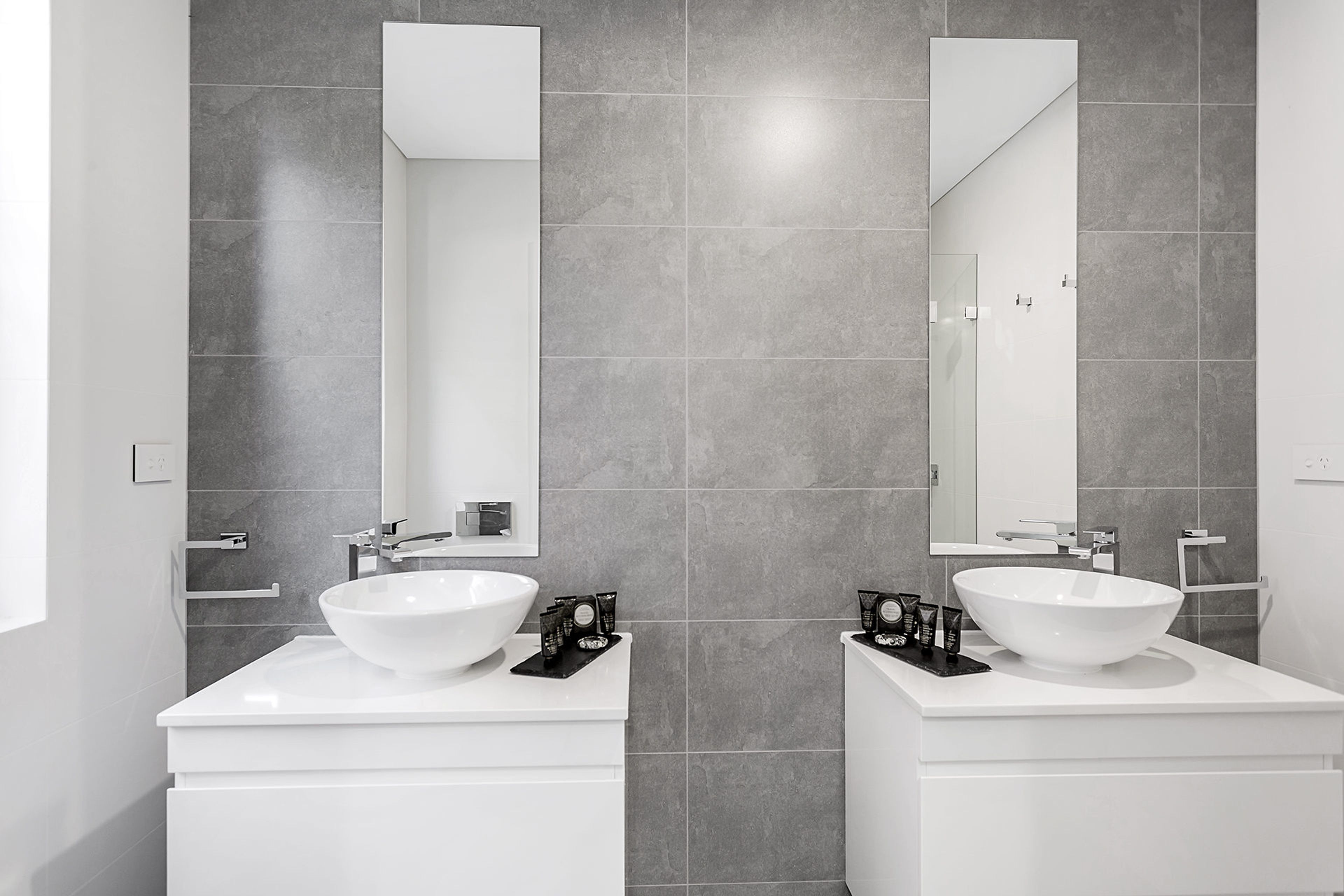 A modern bathroom with two white sinks, large mirrors, and grey tiled walls.