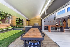 Un espace extérieur aménagé pour les repas, avec une longue table et des chaises en bois, un barbecue et un mur décoré de planches de surf et de cannes à pêche.