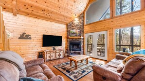 Interior acogedor de una cabaña con paredes de madera, chimenea de piedra, televisor y un ventanal con vistas al bosque.