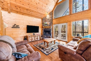 A cozy cabin interior with wooden walls, a stone fireplace, a television, and a large window offering a view of the forest. - Gatlinburg cabin, renovated 3 Brm, Sleeps 8, FP, Pool Table, Hot Tub, WIFI (Gatlinburg)