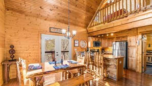 Interior rústico de una cabaña con una mesa de comedor de madera, bancos, una cocina con refrigerador y una zona de altillo con balcón.