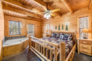 Quarto de cabana de toras com cama de madeira, mesa de cabeceira, abajur, ventilador de teto e uma janela grande.