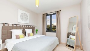 A bedroom with a bed, a bedside table, a lamp, a window with curtains, a wall-mounted mirror, and a ceiling light.