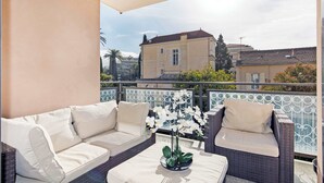 Un balcon avec un canapé en osier et une table, un pot d’orchidée et une vue sur les bâtiments en arrière-plan.