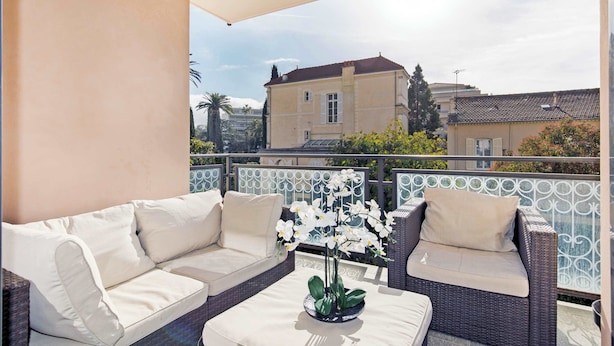 A balcony with a wicker sofa and a table, a potted orchid, and a view of buildings in the background.