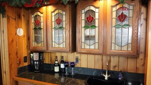 Une cuisine avec des meubles de rangement en bois, un plan de travail noir, une machine à café, des bouteilles de vin et des verres à vin.