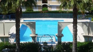 Une piscine où se trouvent deux personnes, entourée de palmiers et, au fond, un bâtiment avec un toit en tuiles.
