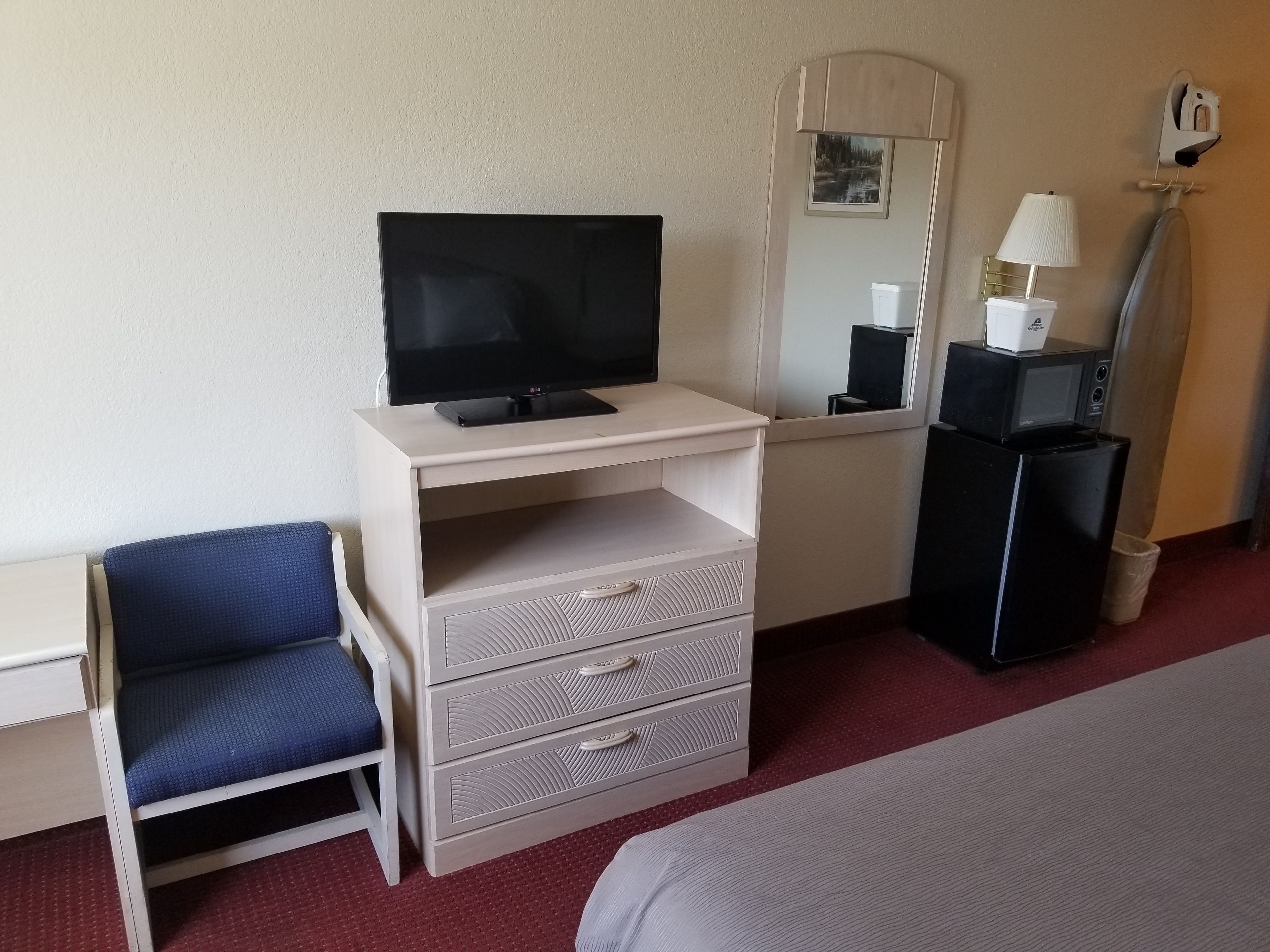 A hotel room with a TV on a desk, a chair, a dresser with drawers, a mirror, a lamp, and a microwave.