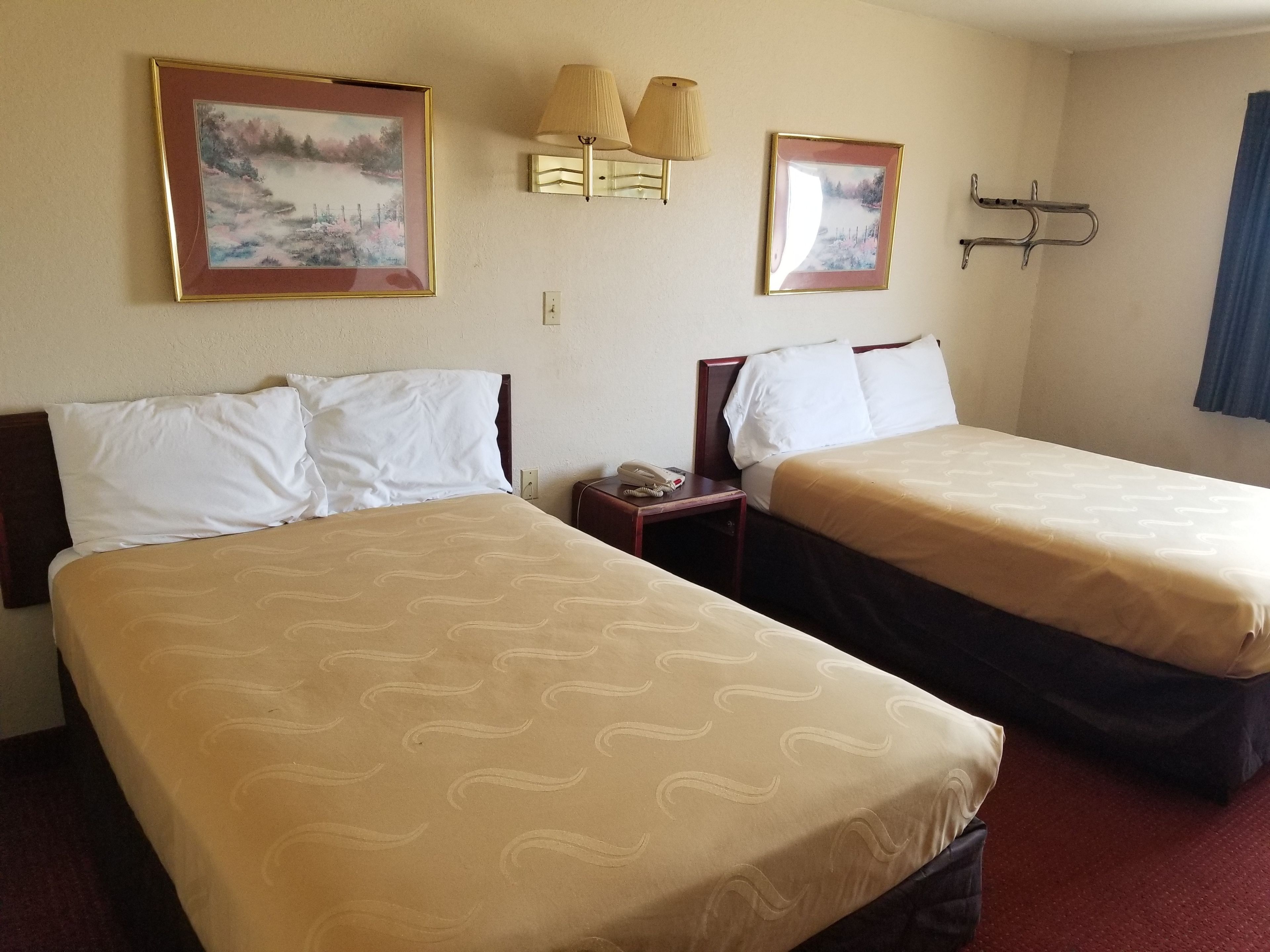 Habitación de hotel con dos camas, cada una con sábanas blancas y un estampado beige. Hay dos lámparas de pared, un cuadro enmarcado y un perchero.