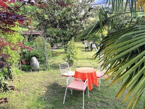 Un área de estar al aire libre con una mesa y sillas, rodeada de abundante vegetación y vista a un edificio al fondo.