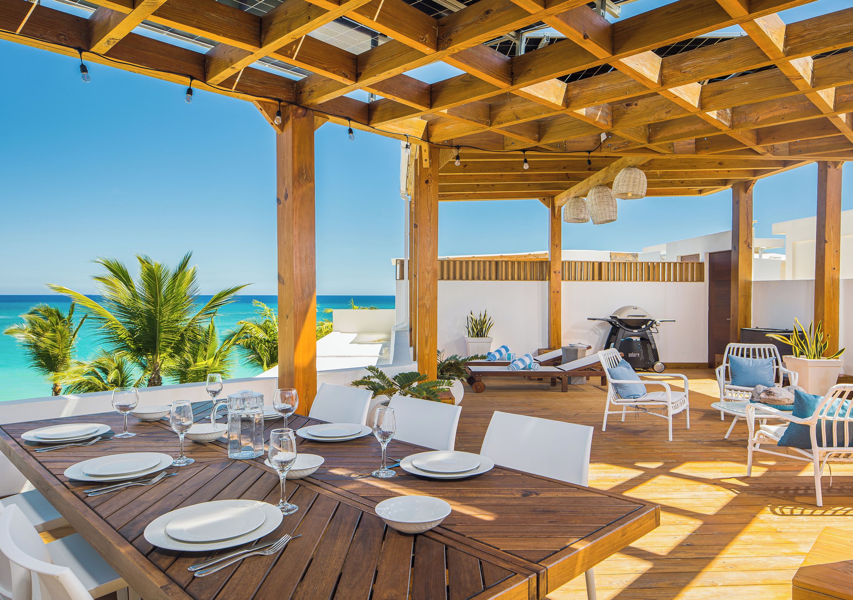 An outdoor dining area with a view of the ocean, wooden pergola, and white furniture.