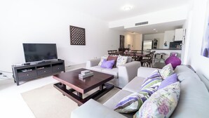 A modern living room with a flat-screen TV, a dark wooden coffee table, and a sofa with colorful pillows. There is a dining area with a table and chairs in the background. - Seawinds Cabarete Beachfront Penthouse, Stunning Balcony Views, Sleeps 6 (Cabarete)