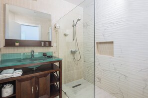 Un baño con cabina de ducha de vidrio, pared con azulejos de mármol, tocador de madera con lavamanos de vidrio y un espejo sobre el tocador.