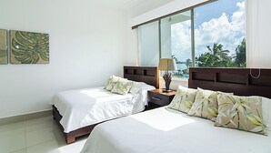 A hotel room with two beds, a nightstand, a lamp, and a large window offering a view of palm trees and the sky. - Seawinds Cabarete Beachfront Penthouse, Stunning Balcony Views, Sleeps 6 (Cabarete)