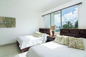 A hotel room with two beds, a nightstand, a lamp, and a large window offering a view of palm trees and the sky. - Seawinds Cabarete Beachfront Penthouse, Stunning Balcony Views, Sleeps 6 (Cabarete)