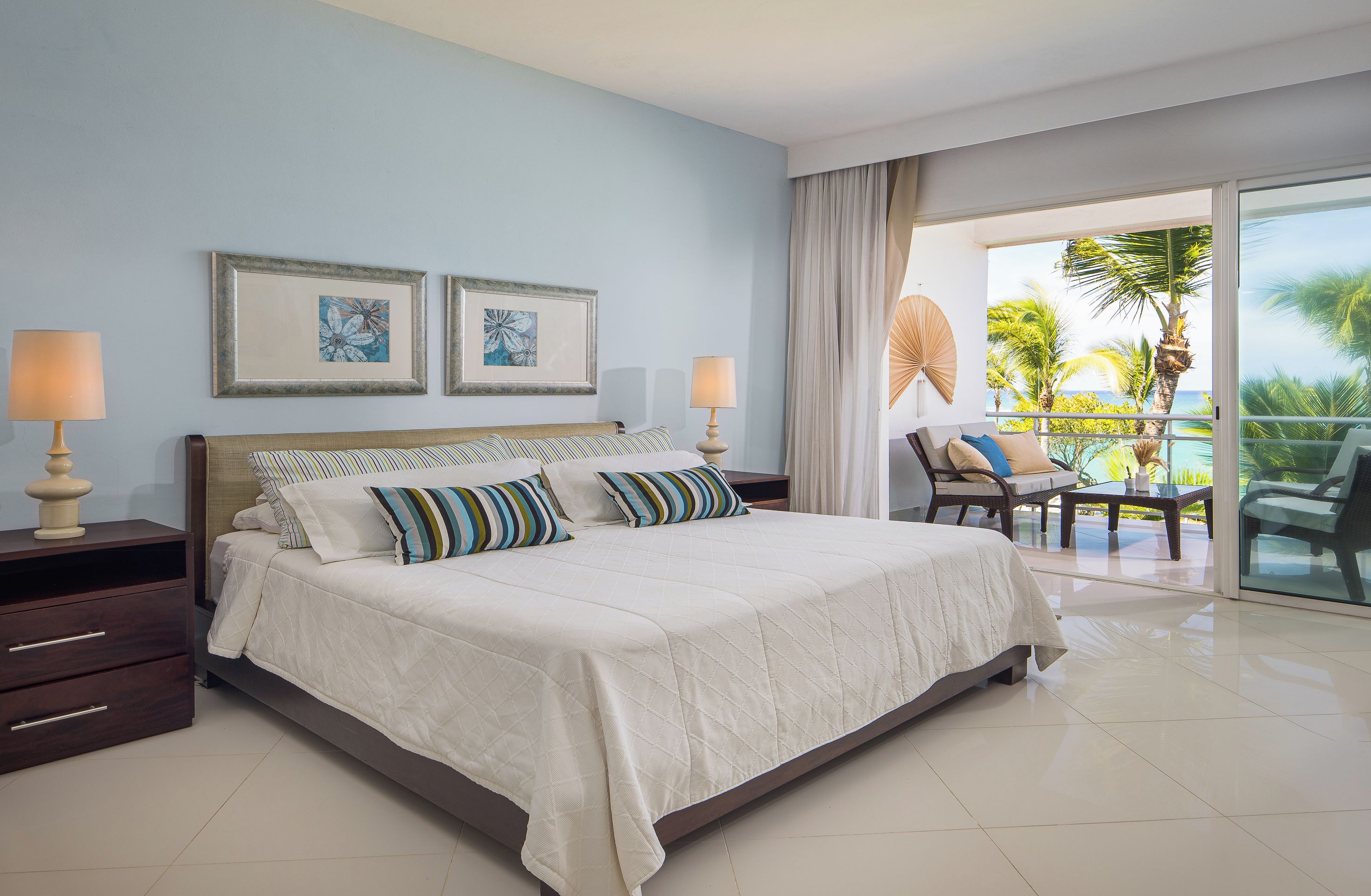 A hotel room with a bed, two bedside lamps, a window with a view of palm trees, and a balcony with outdoor furniture.